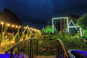 A garden with a metal railing, a building with illuminated geometric shapes, and a clear sky.
