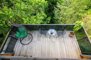 A wooden deck with a glass railing, a green chair, two black chairs, a small round table with a vase, and a potted plant.