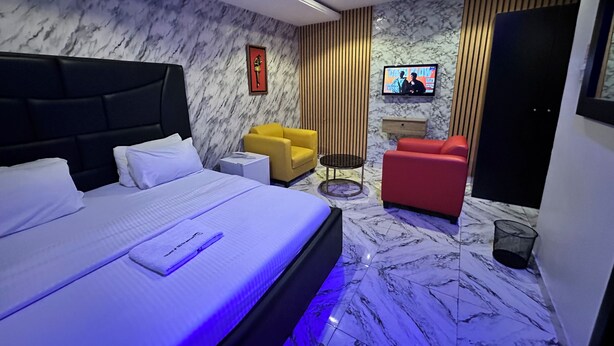 A modern hotel room with a large bed, a TV, a red armchair, a yellow armchair, and a small table. - Desami Hotel (Lagos)