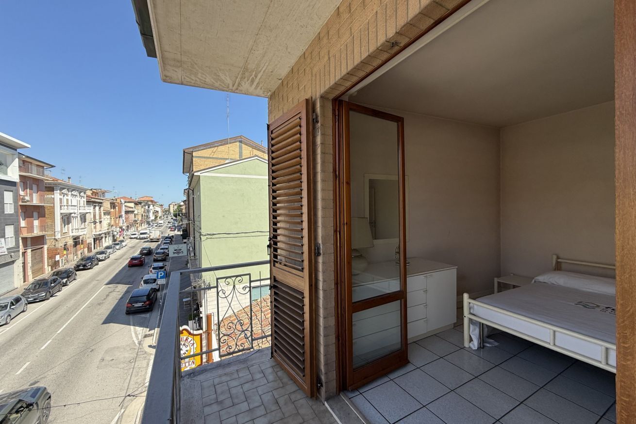 Un balcone con vista sulla strada, una porta scorrevole, un letto e un tavolino.