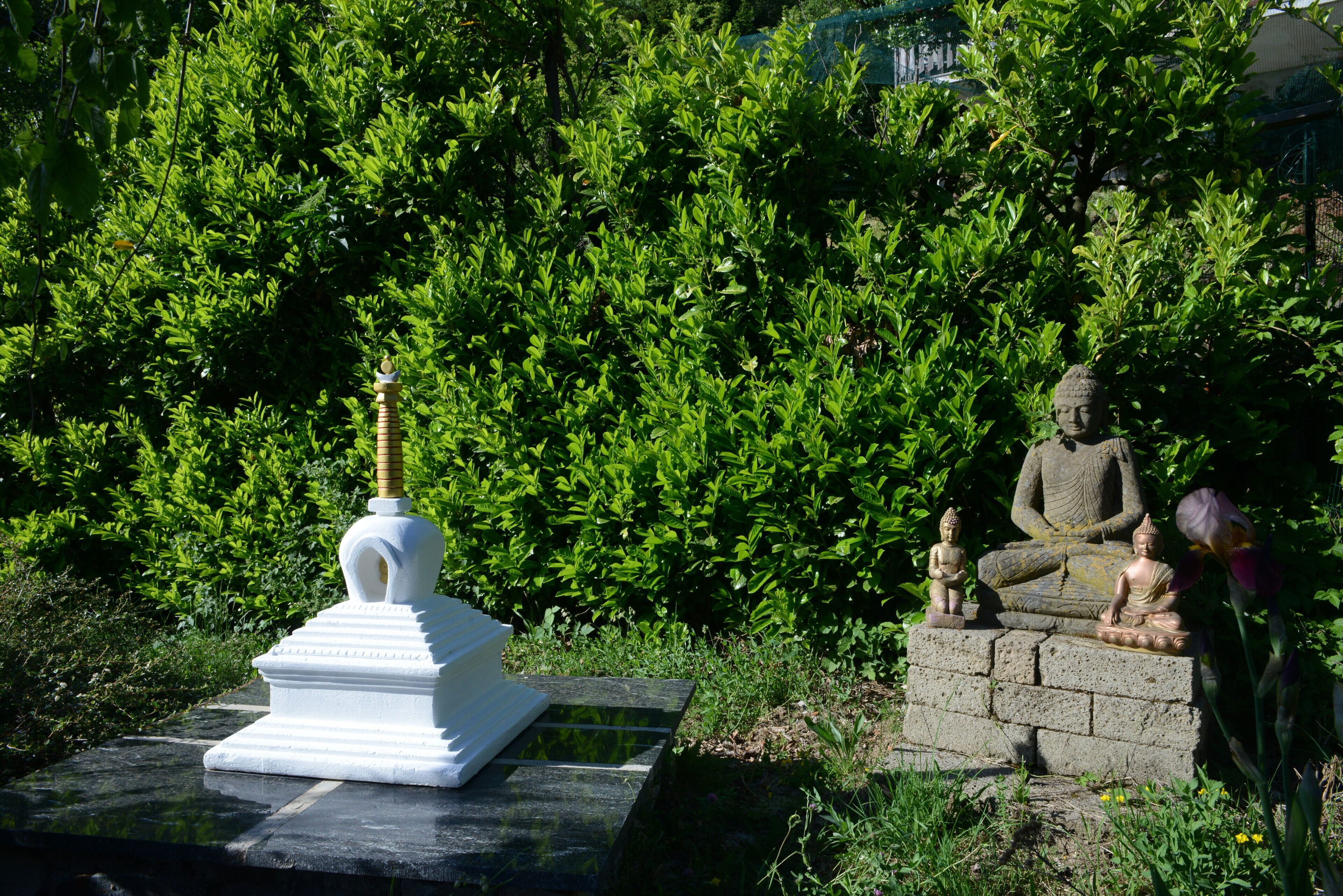 Une statue représentant une figure assise, placée sur un socle en pierre et entourée d’une végétation luxuriante.