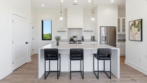 Una cucina moderna con isola centrale, tre sgabelli da bar, illuminazione a sospensione e frigorifero in acciaio inossidabile.