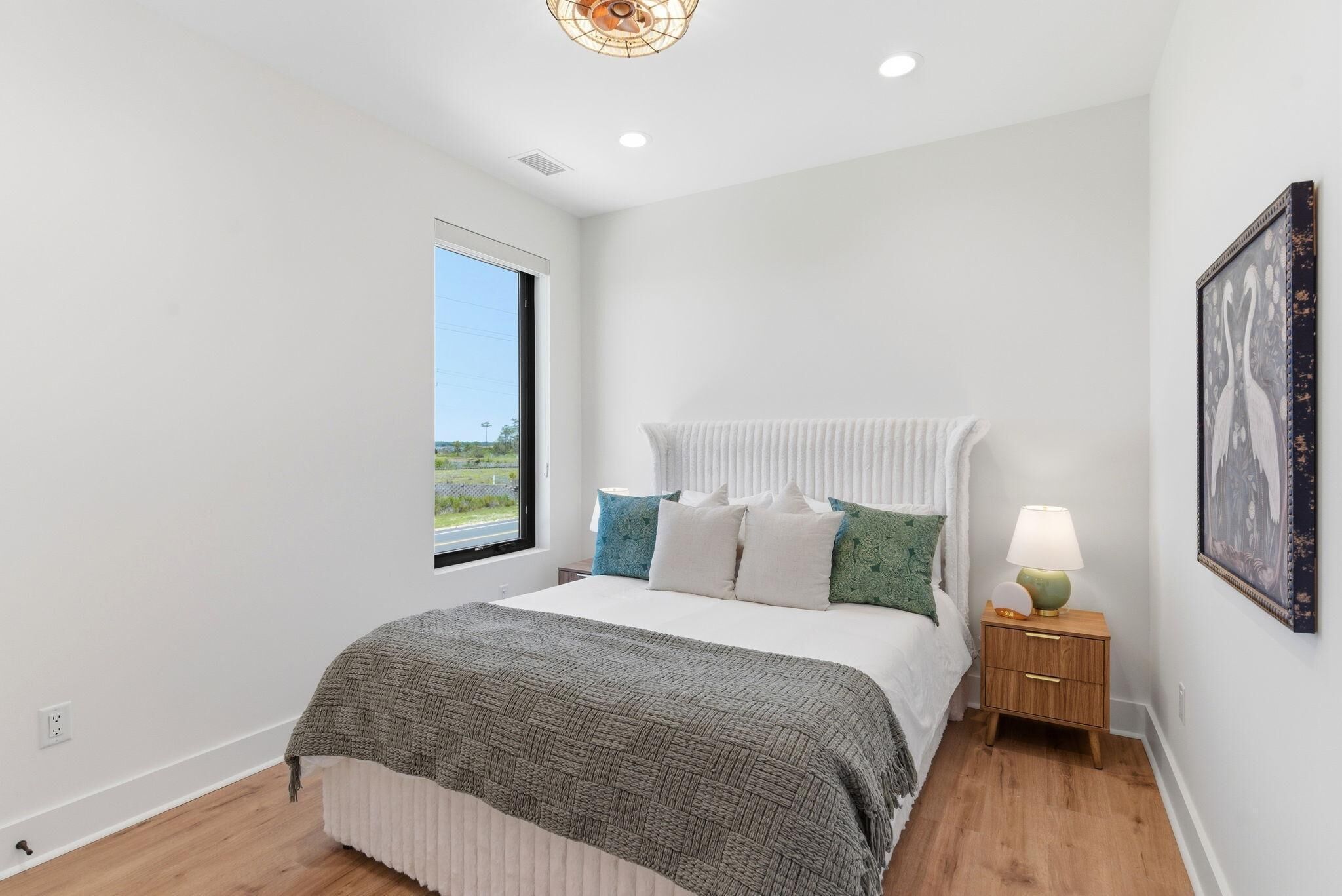 A bedroom with a large window, a bed with a white headboard, a bedside table with a lamp, and a framed picture on the wall.