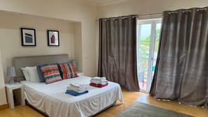 Ein Schlafzimmer mit einem Bett, Kissen, einem Nachttisch, einer Lampe und Blick durch ein Fenster mit Vorhängen.