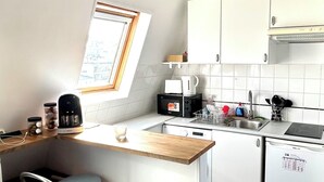 Kompakti keittiö, jossa on puinen työtaso, kaksi sinistä baarijakkaraa, kahvinkeitin, vedenkeitin, astianpesukone ja ikkuna, josta on näkymä.