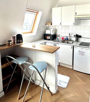 A compact kitchen with a wooden countertop, two blue bar stools, a coffee maker, a kettle, a dishwasher, and a window with a view. - Charming studio apartment in Paris with a balcony, right near lines 1, 2, 6, 9, and the RER A! (Paris)