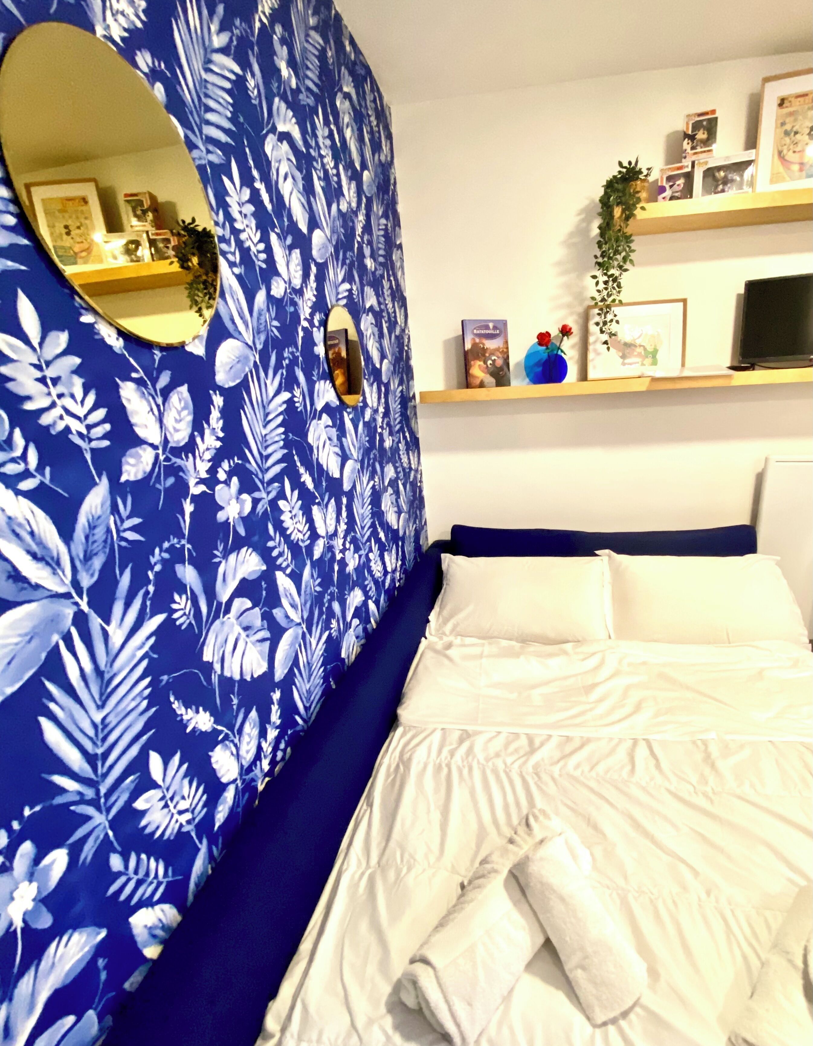 A bedroom with a floral-patterned headboard, a bed with white bedding, a mirror, and a shelf with various items.