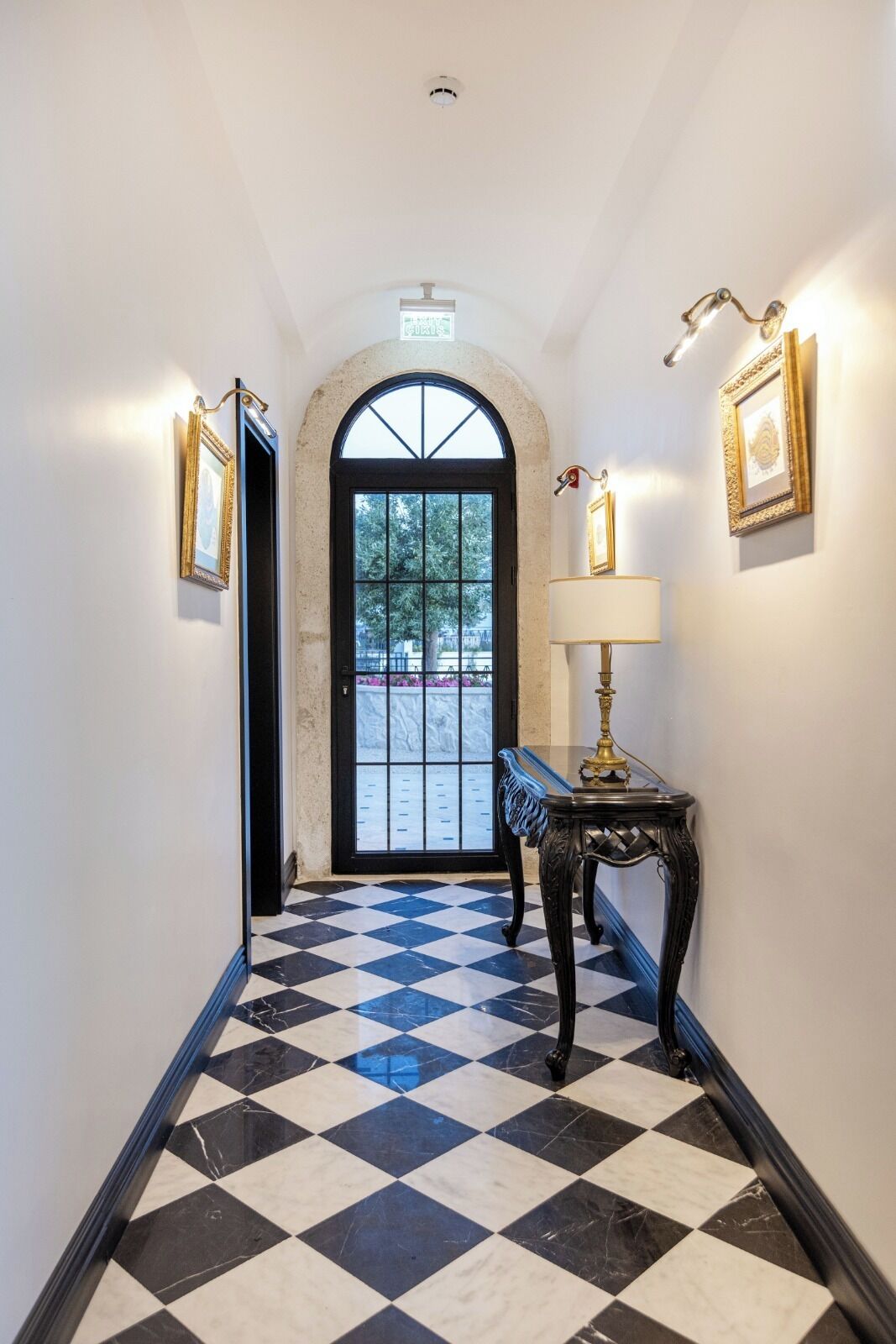 A hallway with a checkered floor, a black door with a glass panel, and wall-mounted lamps.