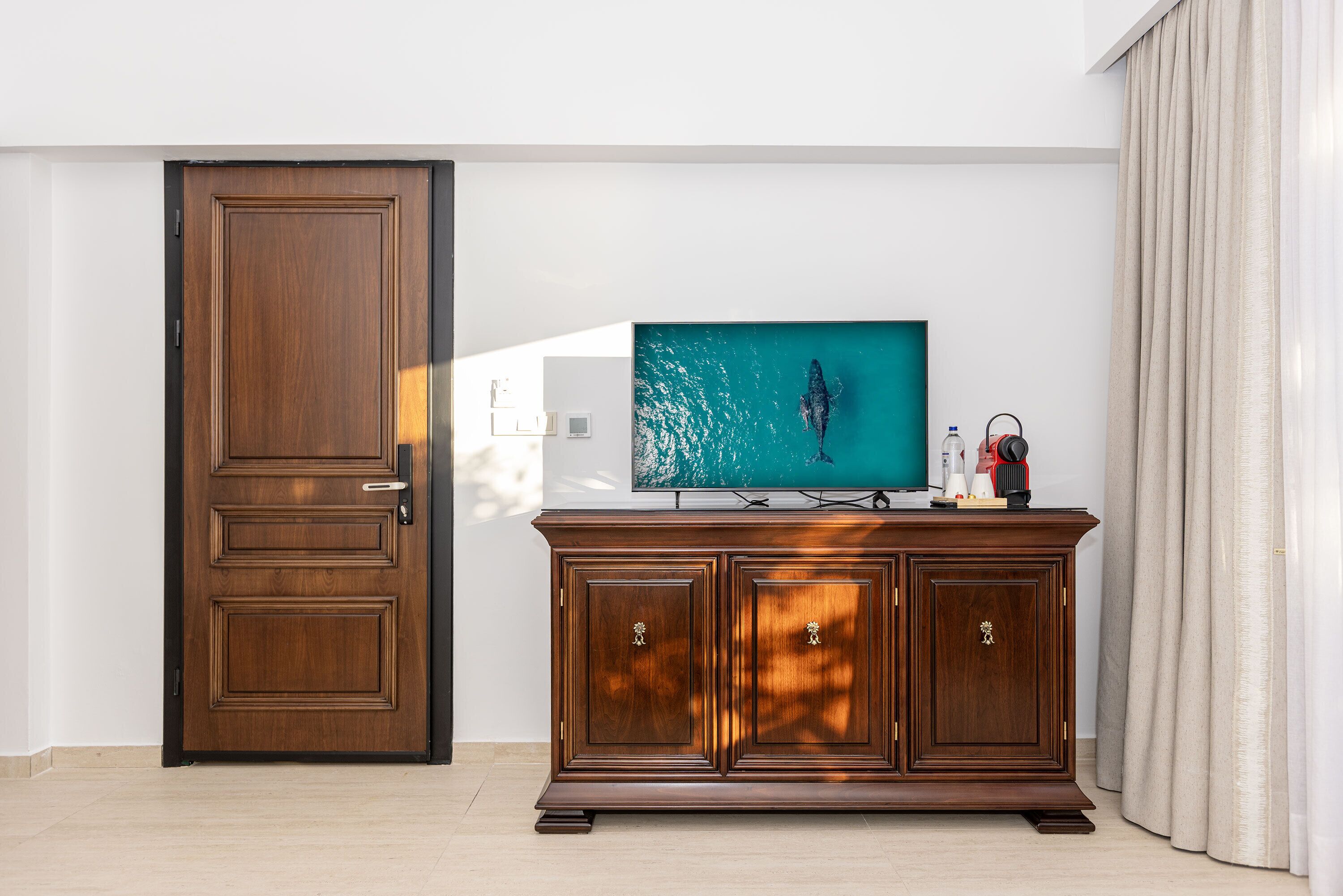 Ein Zimmer mit Holztür, einem Fernseher auf einem Holztisch und einem großen Fenster mit Vorhängen.