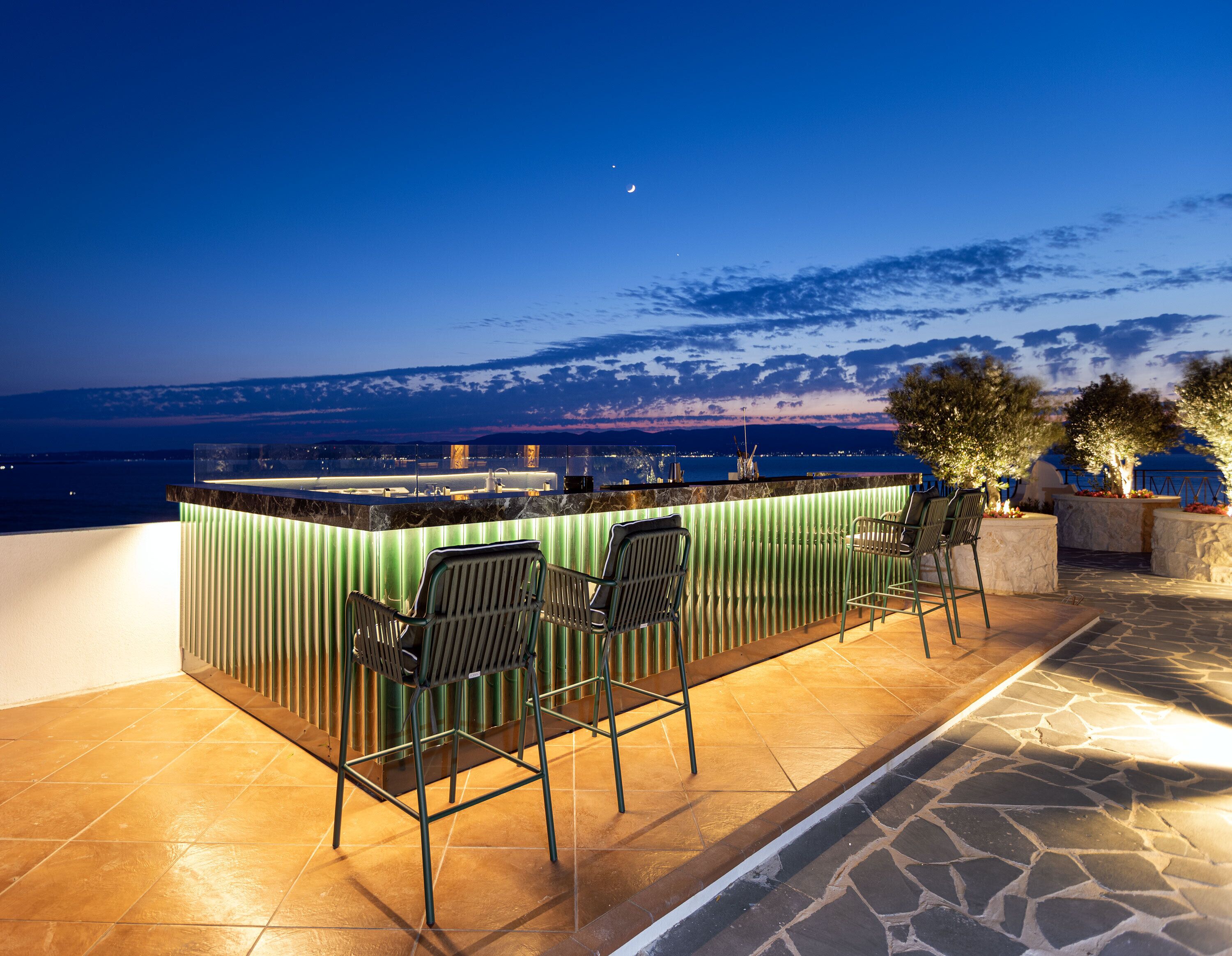 An outdoor bar area with a view of the sea and mountains at dusk.