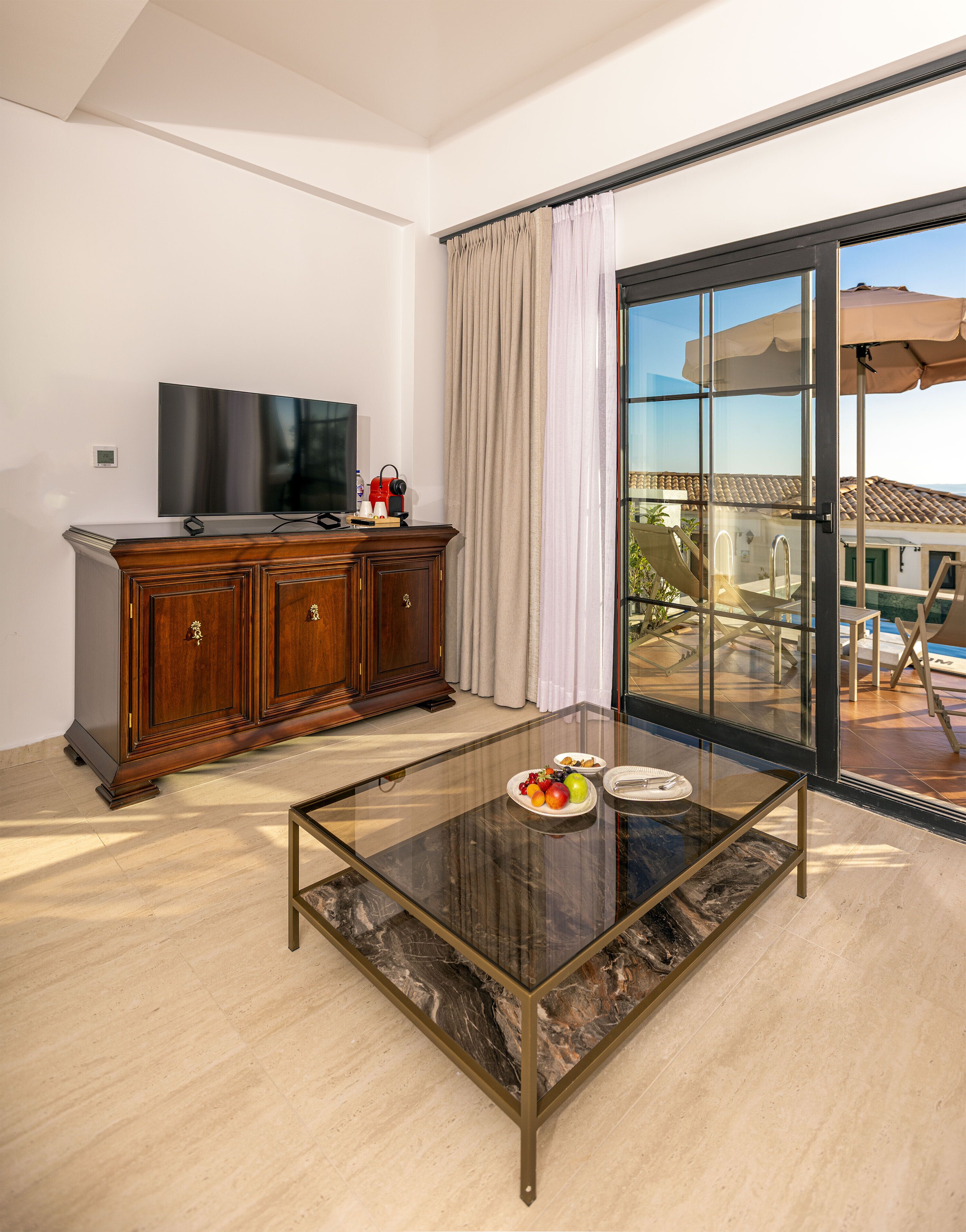 A modern living room with a glass coffee table, wooden cabinet, flat-screen TV, and a view of a pool and outdoor area.