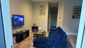 A modern living room with a sectional sofa, a flat-screen TV, a small side table, and a decorative wall piece. - Charming 3-bedroom 2 bath house in lively Houston (Houston)