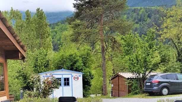 Une terrasse en bois avec des meubles d’extérieur, un petit bâtiment bleu et blanc portant l’inscription « 10 », et une voiture garée à proximité.