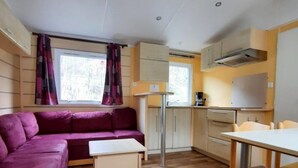 A compact living space with a sofa, a small table, a kitchenette, and a window with curtains. - Mobile home with terrace and TV - 6 guests (Annot)