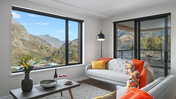 Un salon moderne avec un canapé, une table basse et un vase de fleurs. Une grande fenêtre offre une vue sur les montagnes et une vallée.