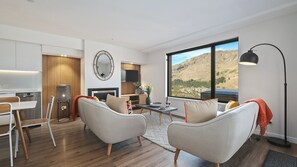 A modern living room with a dining area, a fireplace, and a large window offering a mountain view. - Shotover River Hideaway - 1 Bed (Queenstown)