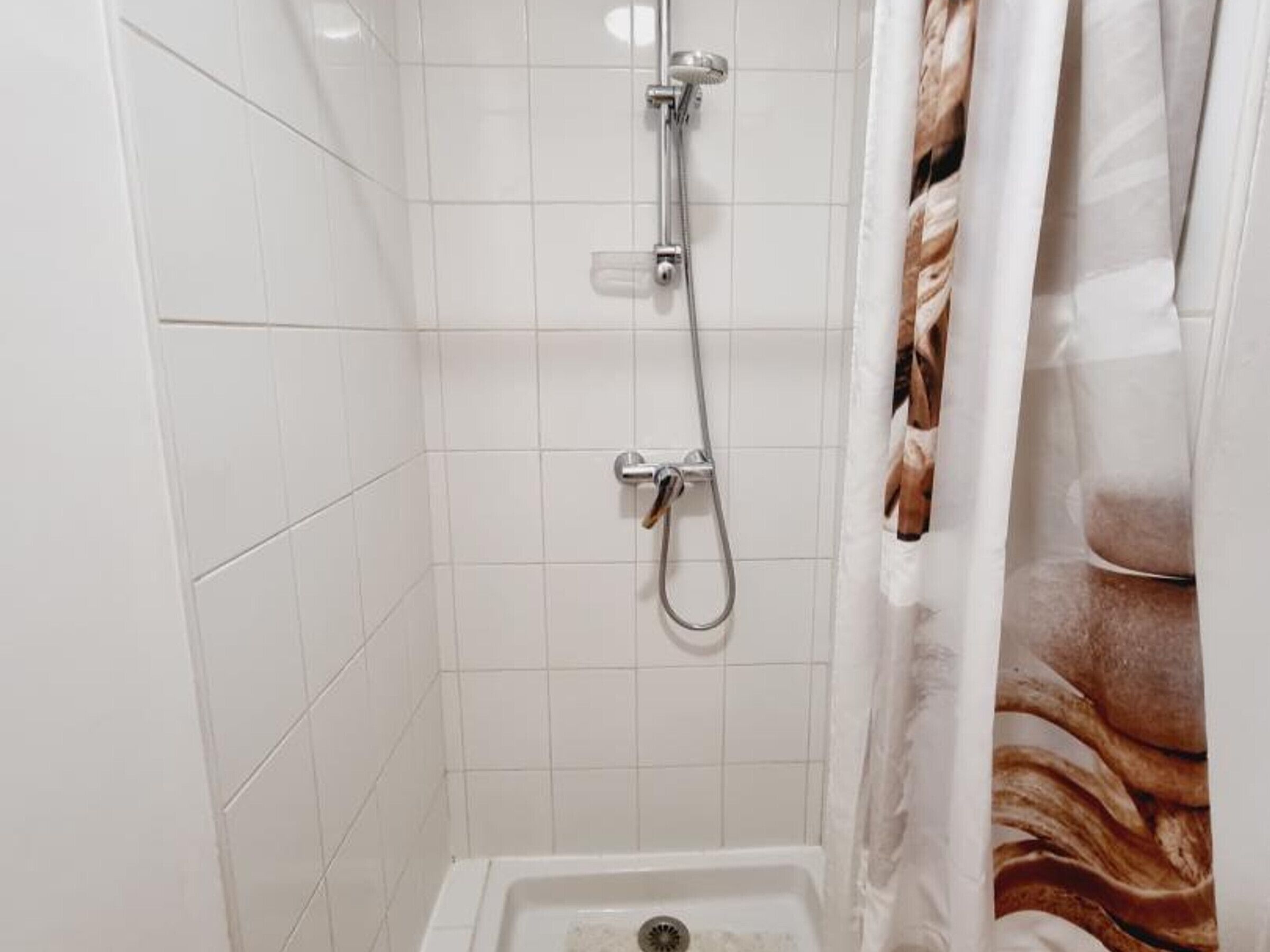 A shower with white tiles, a showerhead, and a curtain with a brown pattern.