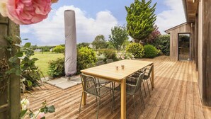 Una terrassa de fusta amb taula i cadires, envoltada de vegetació i un cel serè.