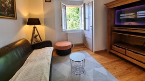 Een woonkamer met een bank, een lamp, een klein rond tafeltje, een televisie en een raam met uitzicht op groen.