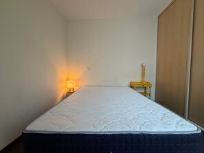 Un lit simple avec un matelas blanc et une couverture matelassée de couleur foncée, dans une pièce aux murs unis et dotée d’une armoire en bois.