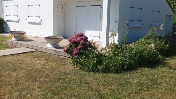 Ein weißes Gebäude mit Satteldach, einer überdachten Veranda und einem Blumenbeet mit rosa Blüten.