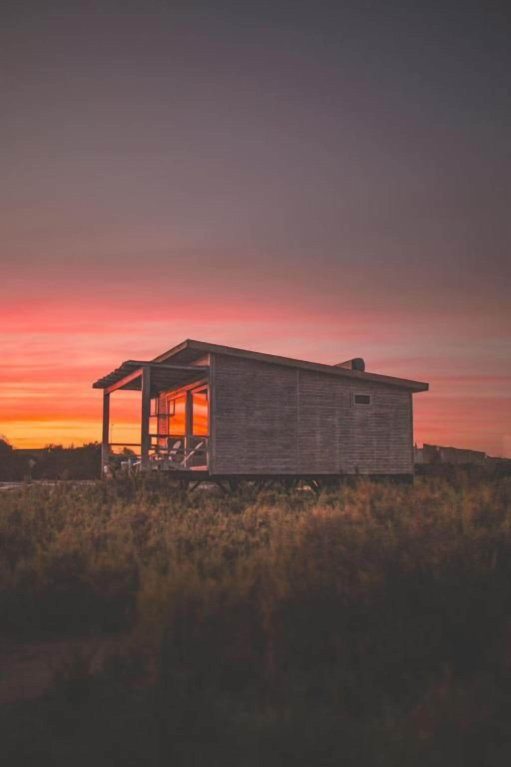 Uma cabana de madeira térrea com varanda, cercada por grama alta, ao pôr do sol.