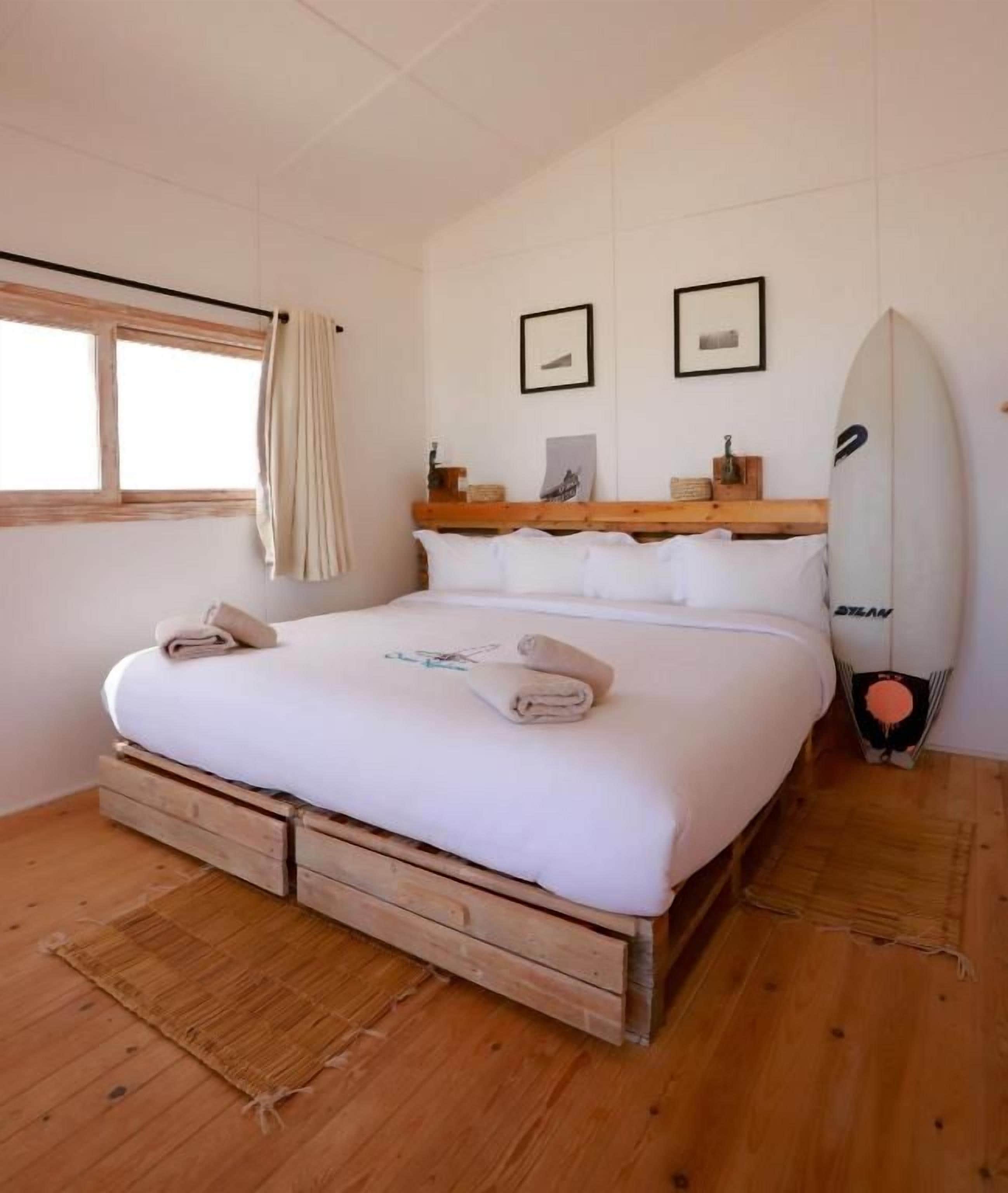 Quarto com cama, piso de madeira, uma prancha de surf e janela com cortinas.