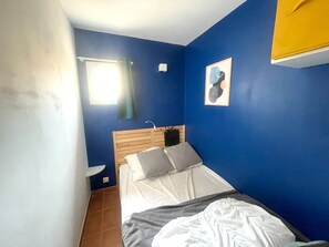 Ein Schlafzimmer mit hölzernem Kopfteil, einem Bett mit weißer Bettwäsche, einem kleinen Nachttisch, einem Fenster mit Vorhang und einer Wandleuchte.