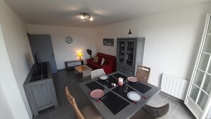 Ein Essbereich mit einem Tisch für vier Personen, umgeben von Korbstühlen. Im Hintergrund befindet sich eine rote Couch und ein Schrank.