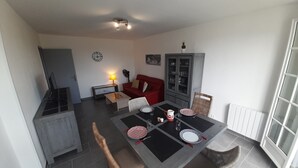 A dining area with a table set for four, surrounded by wicker chairs. A red sofa and a cabinet with glass doors are visible in the background. - Charming T2 city center - 2 guests (La Roche-Posay)