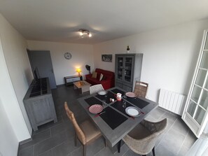 A dining area with a table set for four, surrounded by wicker chairs. A red sofa and a cabinet with glass doors are visible in the background. - Charming T2 city center - 2 guests (La Roche-Posay)