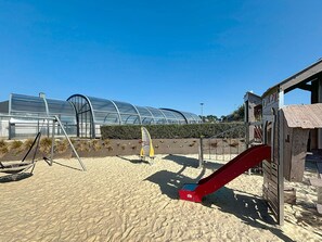 A sandy beach with a swing set, a red slide, and a greenhouse structure in the background. - City mobile home, pets allowed - 4 guests (Jullouville)