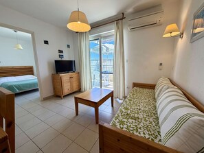 Ein Zimmer mit einem Bett, einem Fernseher auf einer Holzkassette, einer Couch, einem Tisch und einem Balkon mit Vorhängen.