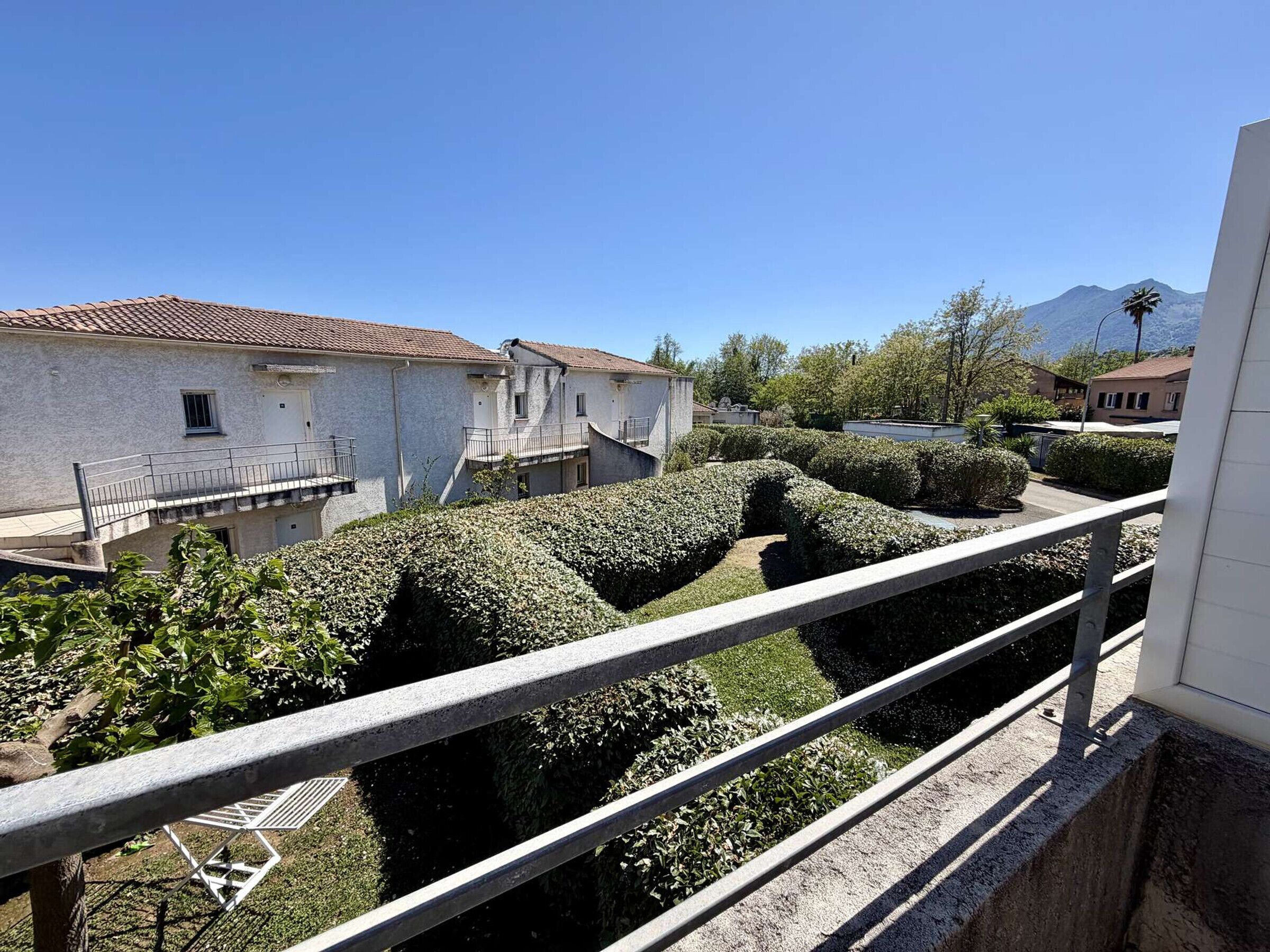 Une cour avec des haies soigneusement taillées, un bâtiment blanc surmonté d’un toit de tuiles et un ciel d’un bleu limpide.