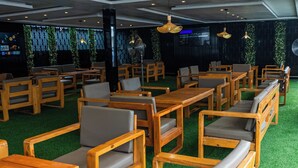 Restaurante com mesas e cadeiras de madeira, piso de grama artificial verde e luminárias pendentes.