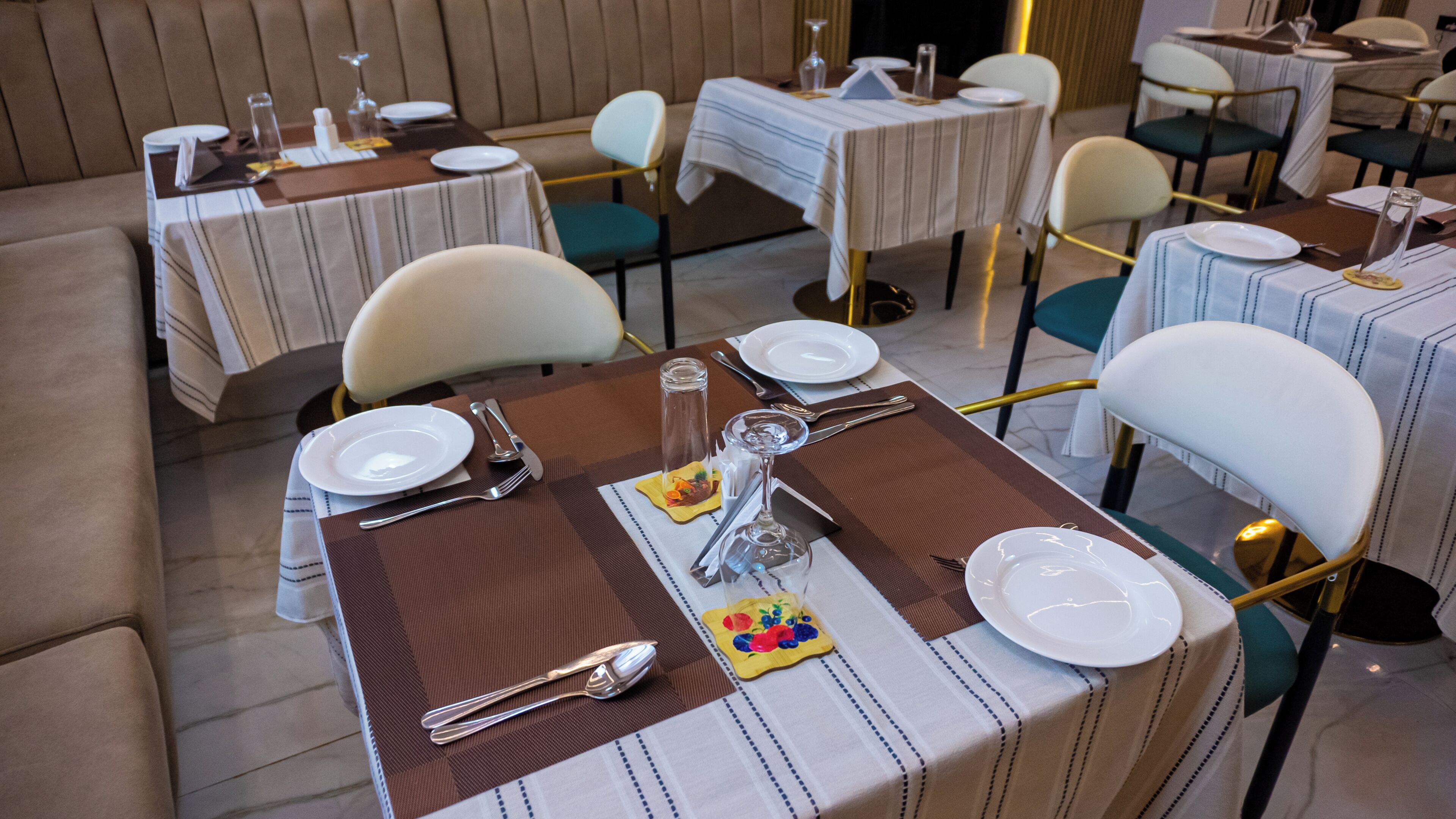 A dining area with tables set for two, featuring striped tablecloths, white plates, cutlery, and a glass of water.