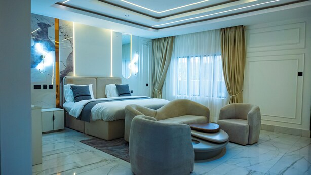 Quarto moderno com uma cama grande, duas poltronas, uma mesinha e chão de mármore.