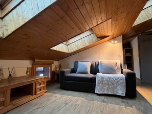 Ein gemütliches Schlafzimmer im Loft-Stil mit schrägem Dach, einer dunkelgrauen Couch, einem Holznachttisch und einem kleinen Tisch.