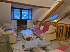 A loft-style living room with a TV, sofa, ottoman, and a view of the city through large windows. - Duplex apartment for 8/10 pers, 150m from cable car (Cauterets)