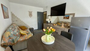 Ein modernes Wohnzimmer mit einer Couch, einem Esstisch mit einer Tulpenvase, einer Küchenzeile mit Kühlschrank und einem an der Wand befestigten Flachbildfernseher.
