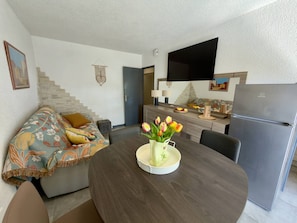 A modern living room with a sofa, a dining table with a vase of tulips, a kitchen area with a refrigerator, and a flat-screen TV mounted on the wall. - Comfortable apartment near the beach - 4 guests (Argelès-sur-Mer)