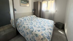 Ein Schlafzimmer mit Bett, einer Tür, einem Fenster mit Vorhängen, einem Nachttisch und einem an der Wand hängenden Bild.