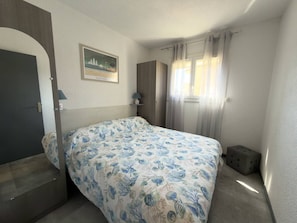 A bedroom with a bed, a door, a window with curtains, a bedside table, and a wall-mounted picture. - Comfortable apartment near the beach - 4 guests (Argelès-sur-Mer)