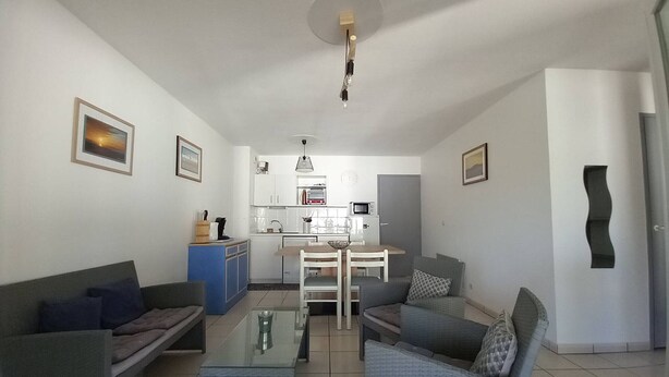 Uma sala de estar com sofá, poltrona e mesa de centro, uma cozinha compacta com micro-ondas, e um espaço de jantar com mesa e cadeiras.