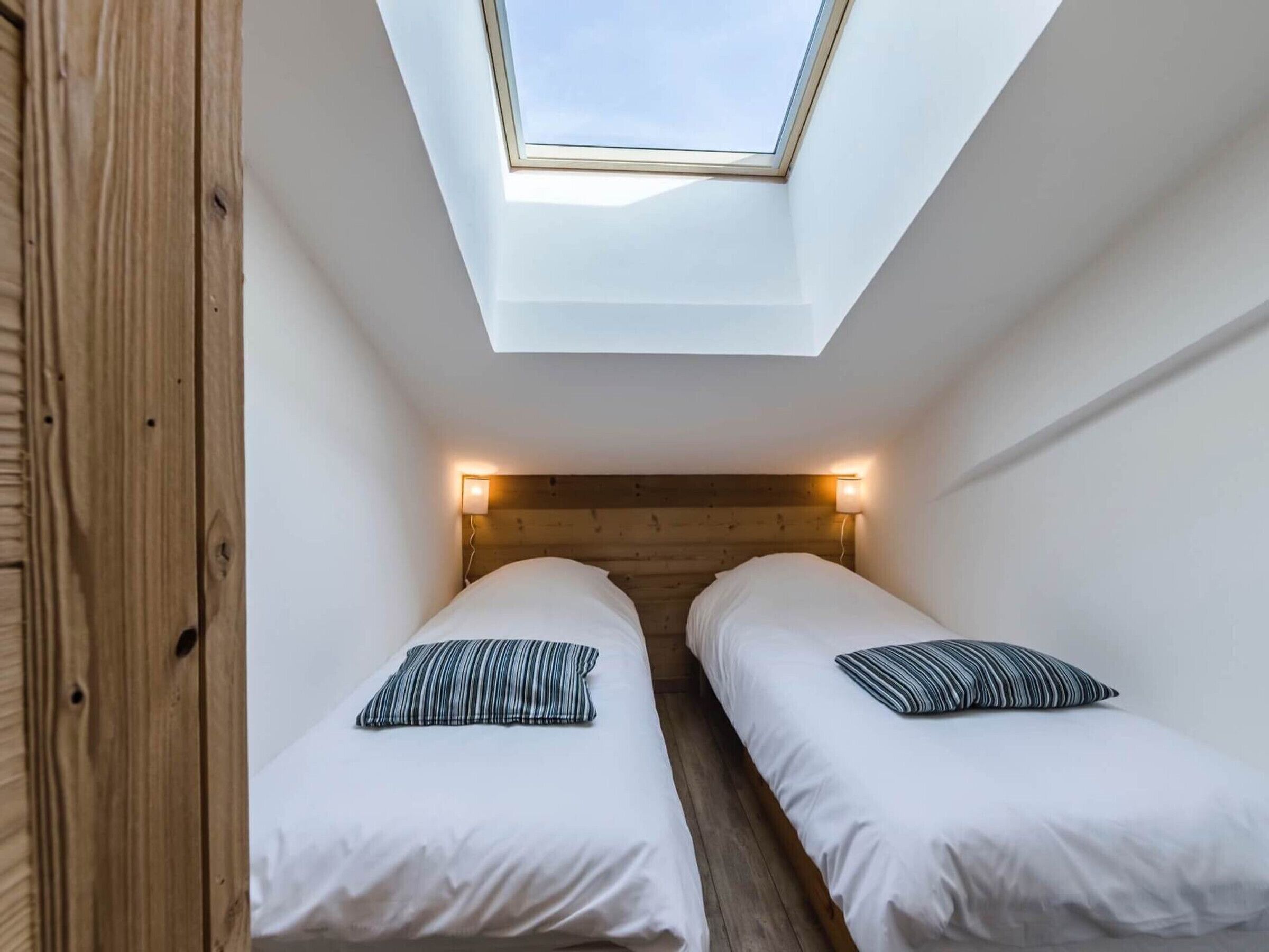 A small, modern bedroom with two beds, a skylight, and wooden headboard.