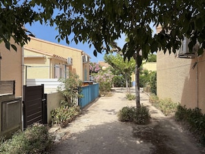 A narrow alleyway with a building on the left, a blue fence, and a tree in the center. - House with AC, near beach - 5 guests (Fleury-Saint-Pierre-la-Mer)