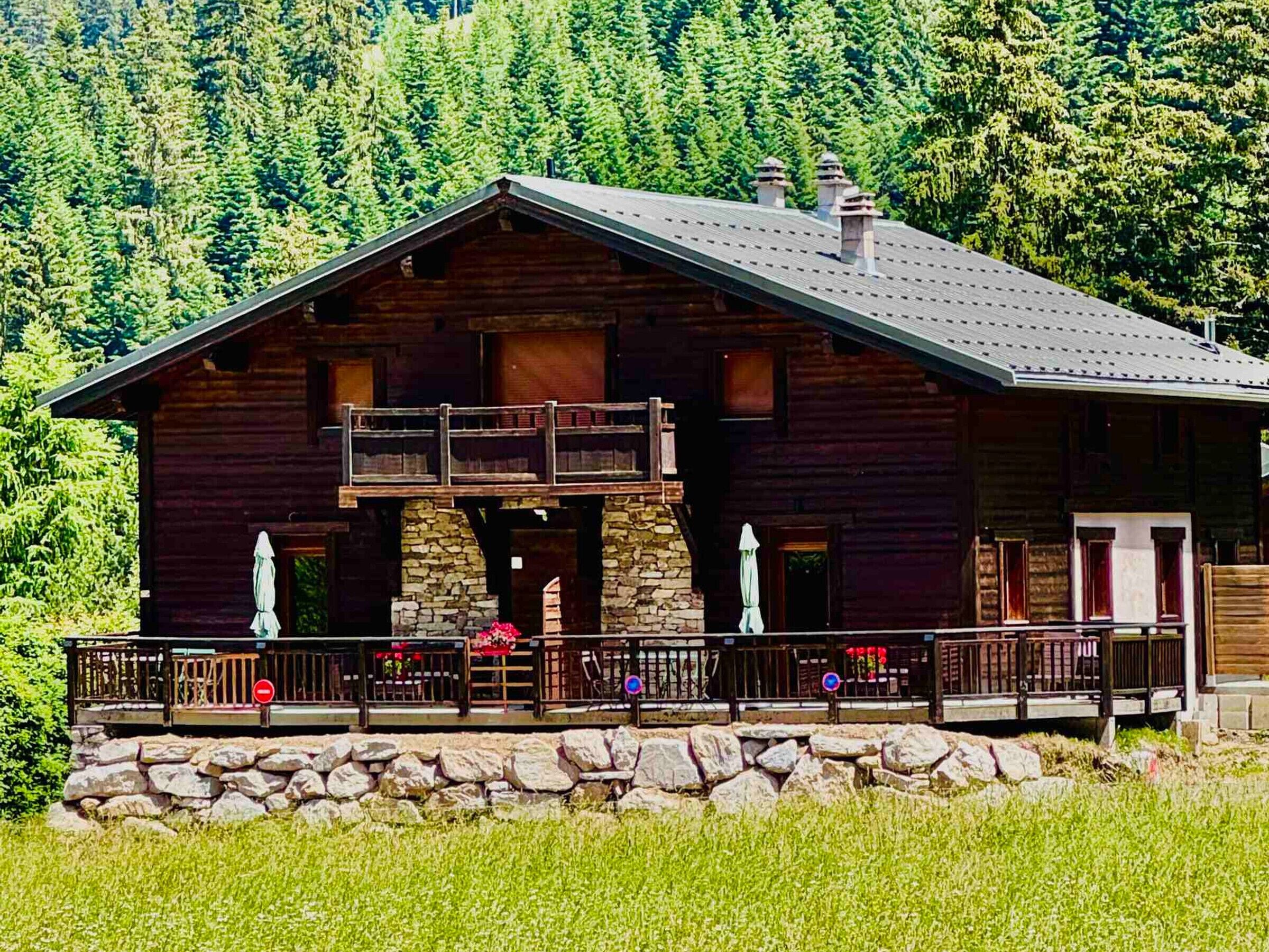 Une cabane en rondins dotée d’un balcon, avec des fondations en pierre et un mur de pierre à l’avant.