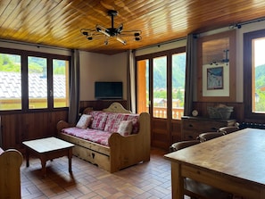 Ein gemütliches Zimmer mit Holzbalkendecke, einer Couch, einem Tisch und großen Fenstern mit Blick ins Freie.
