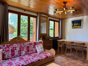 Ein gemütliches Zimmer mit Holzbalkendecke, einer Couch mit gemusterten Kissen, einem Esstisch mit Stühlen und großen Fenstern mit Blick ins Grüne.