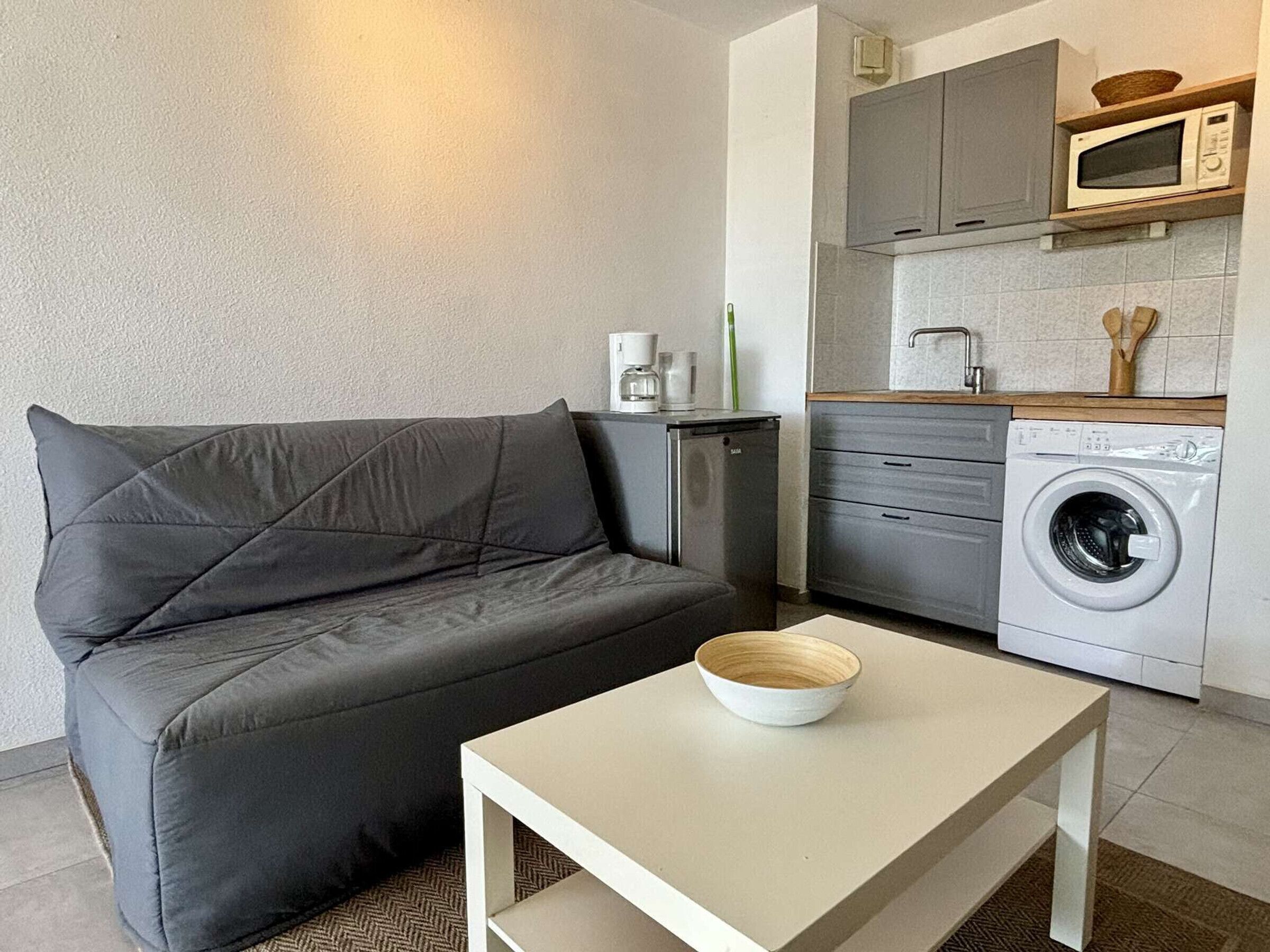A compact living space with a sofa, a small table with a bowl, a washing machine, and a kitchenette with a microwave.